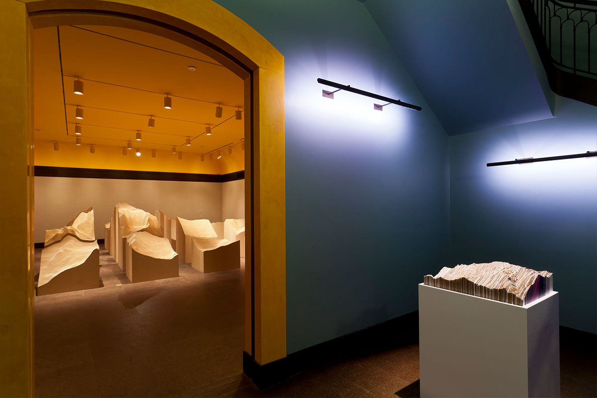 A blue lit gallery featuring a sculptural model on a pedestal; in the background, a yellow lit gallery features a large installation of terrain-like segments of material