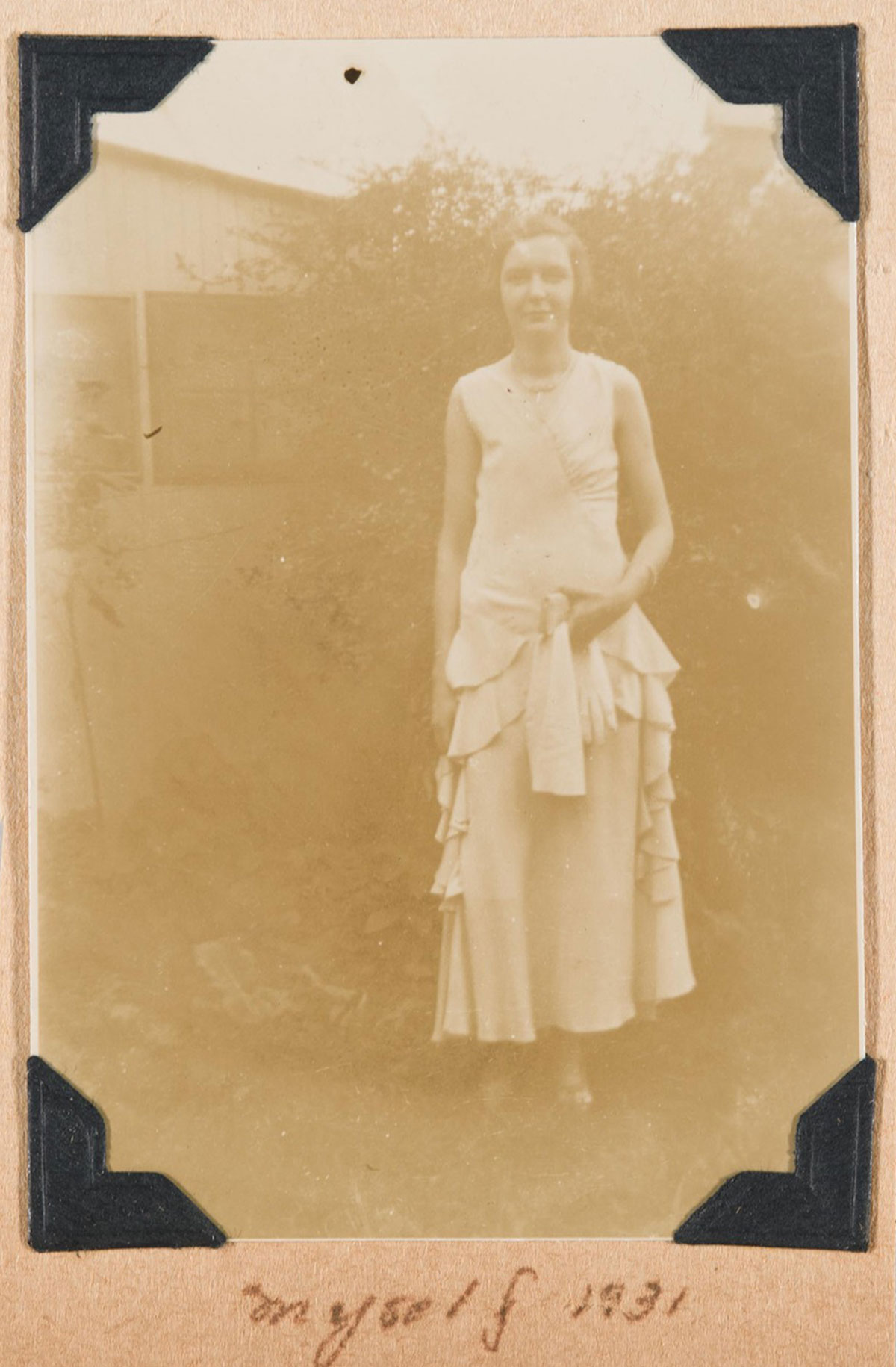A woman wearing a long, light-colored dress stands outside of a house in front of a large bush; underneath the image is written "myself 1931"