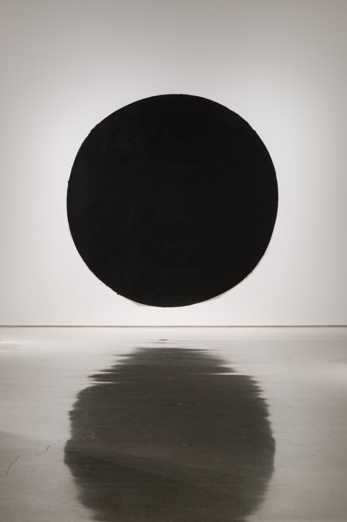 A hand-brushed circle of deep black pigment is tacked directly on a white gallery wall. The circle is reflected in the gallery floor.