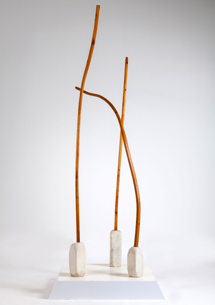 A tall, wooden sculpture with three pieces, each comprised of a narrow, light reddish brown piece of wood reaching up from a short marble vase
