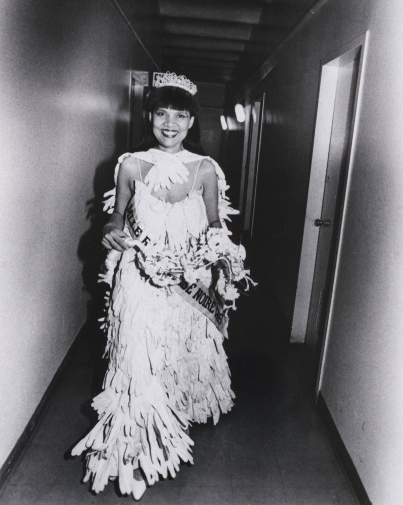 A Black woman wearing a tiara and cap and gown made of long white gloves walks down a narrow hallway, smiling brilliantly at the camera
