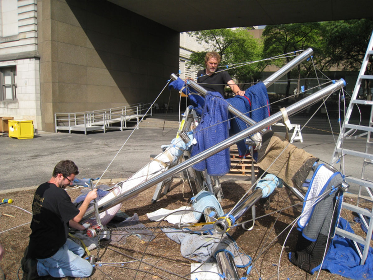A crew of workers install a large, metal sculpture in an outdoor courtyard