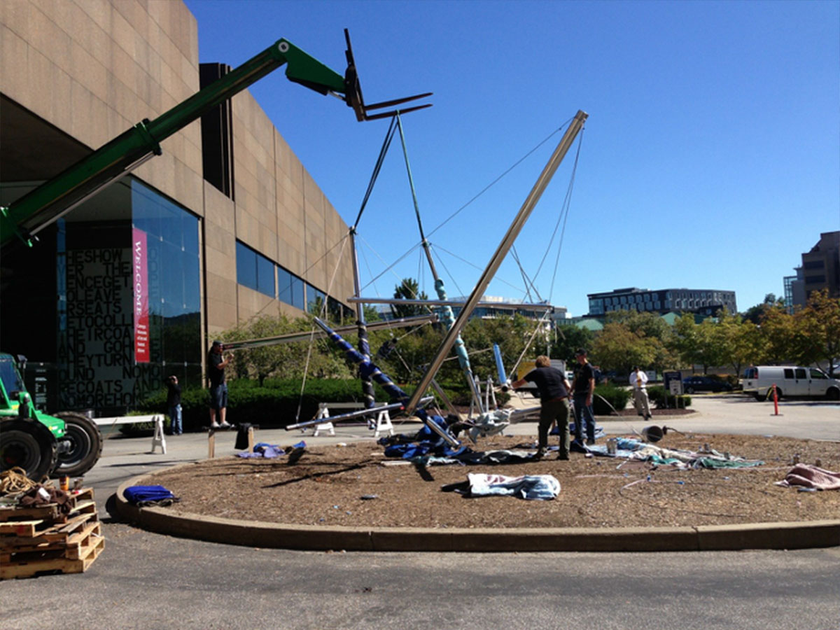 A crew of workers install a large, metal sculpture in an outdoor courtyard