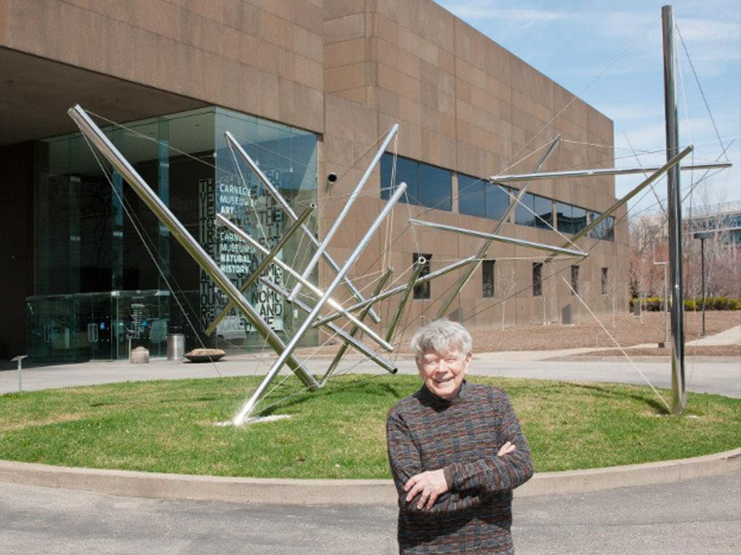 A man with grey hair and a dark sweater stands, smiling, in front of an abstract metal sculpture on a green space in front of a large building