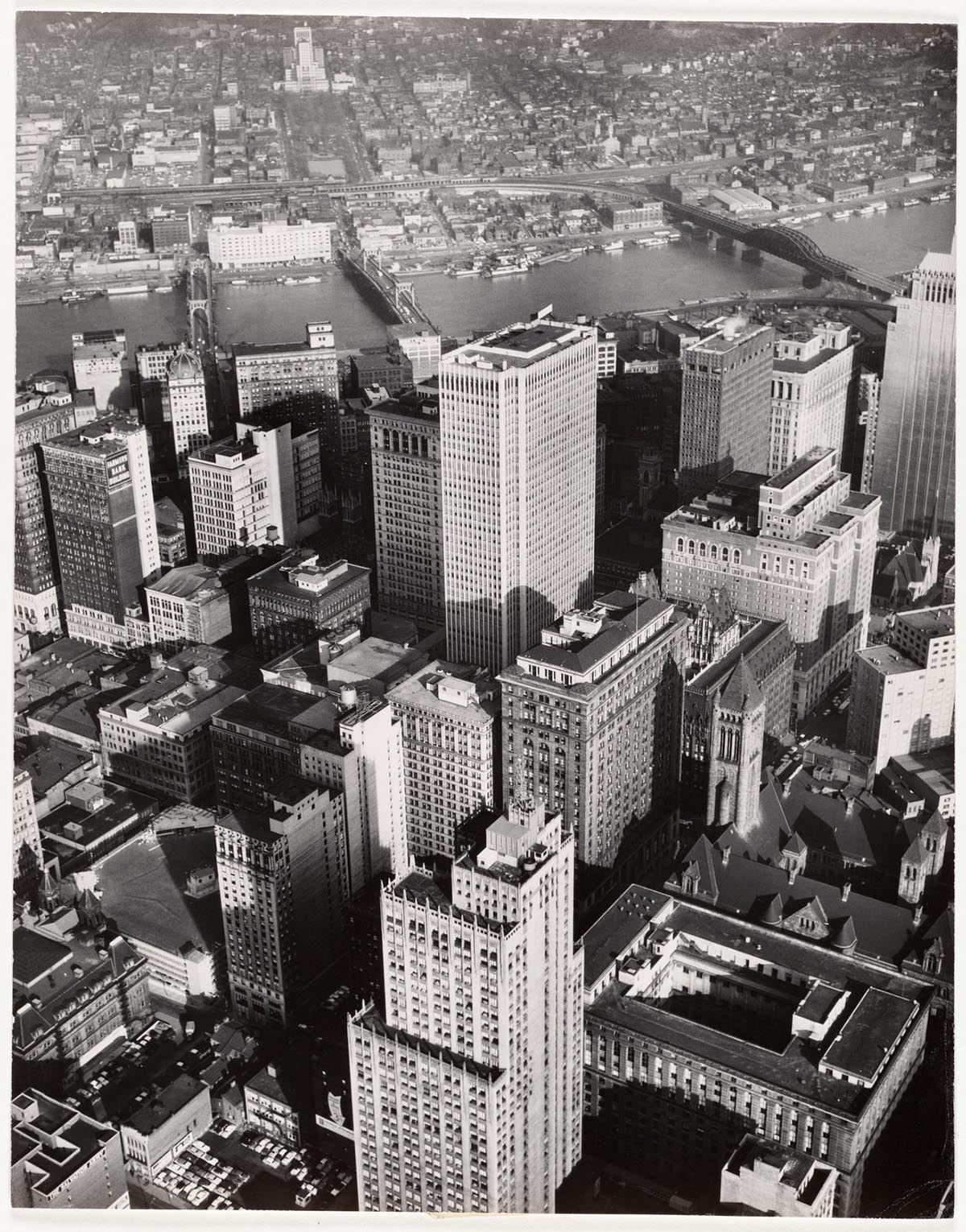 A black-and-white aerial photograph of the city of Pittsburgh looks down on a cluster of skyscrapers.