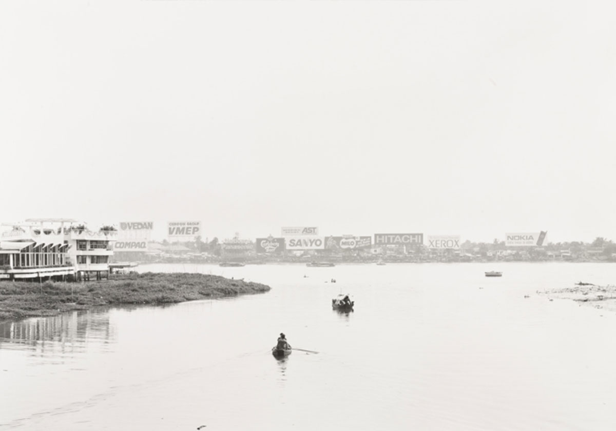 A black and white photo of a wide river with small boats in the center; the far shore of the river features rows of billboards