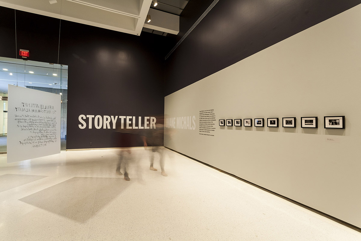 People walk in a large exhibition with framed black and white photos on a wall. Behind them is a black wall with the word "storyteller" in white. A board with illegible text hangs from the wall.