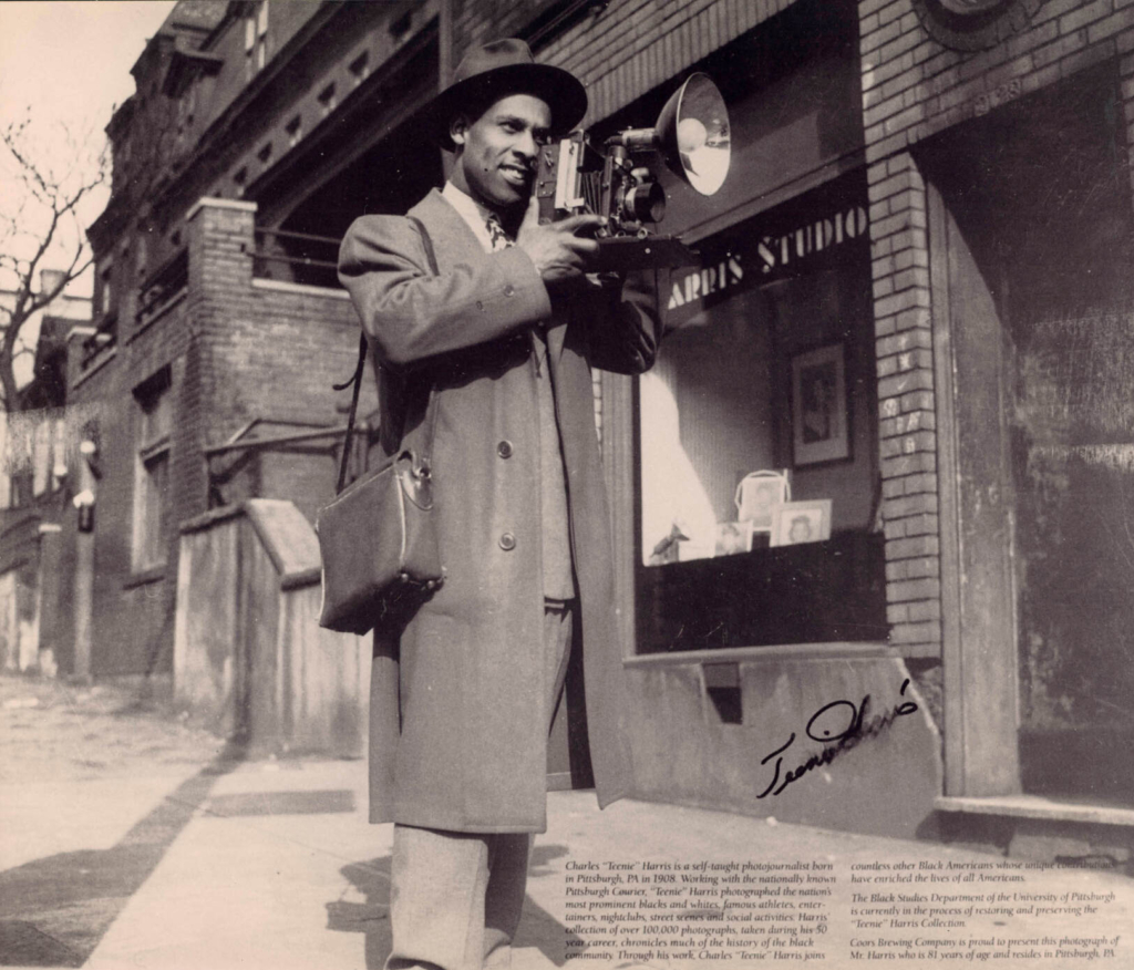 A person in a trench coat and hat stands on a city sidewalk with a camera and large flash in their hand
