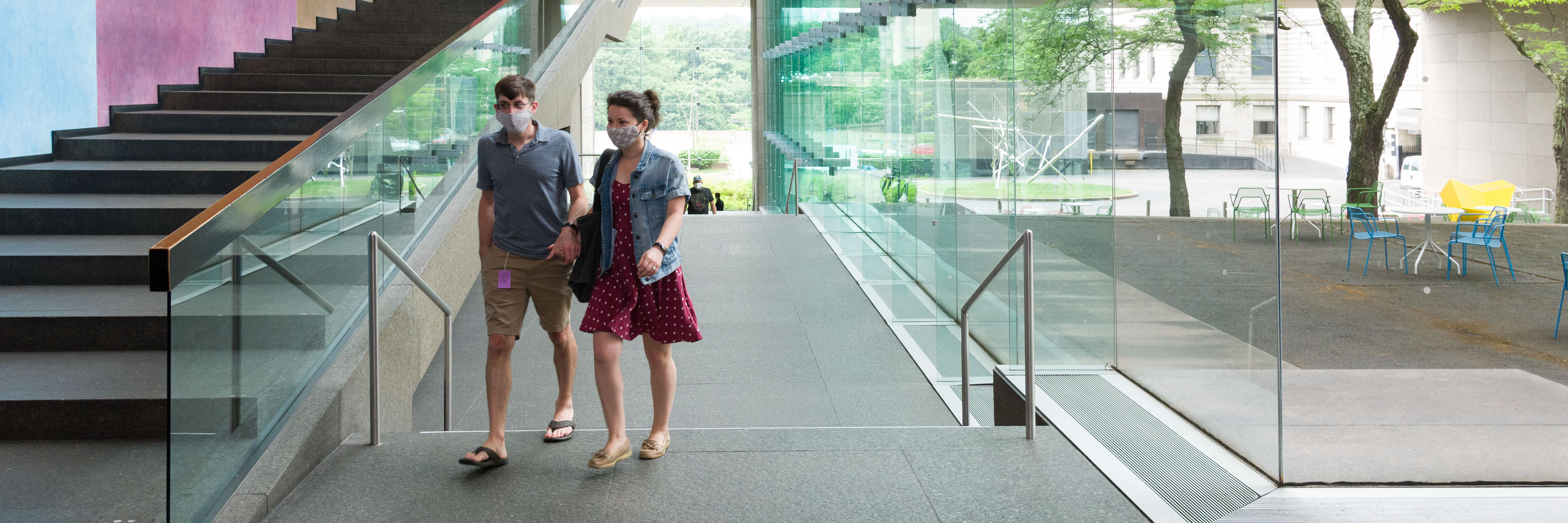 A couple walking past a stairway holding hands with a courtyard visible through glass walls behind them