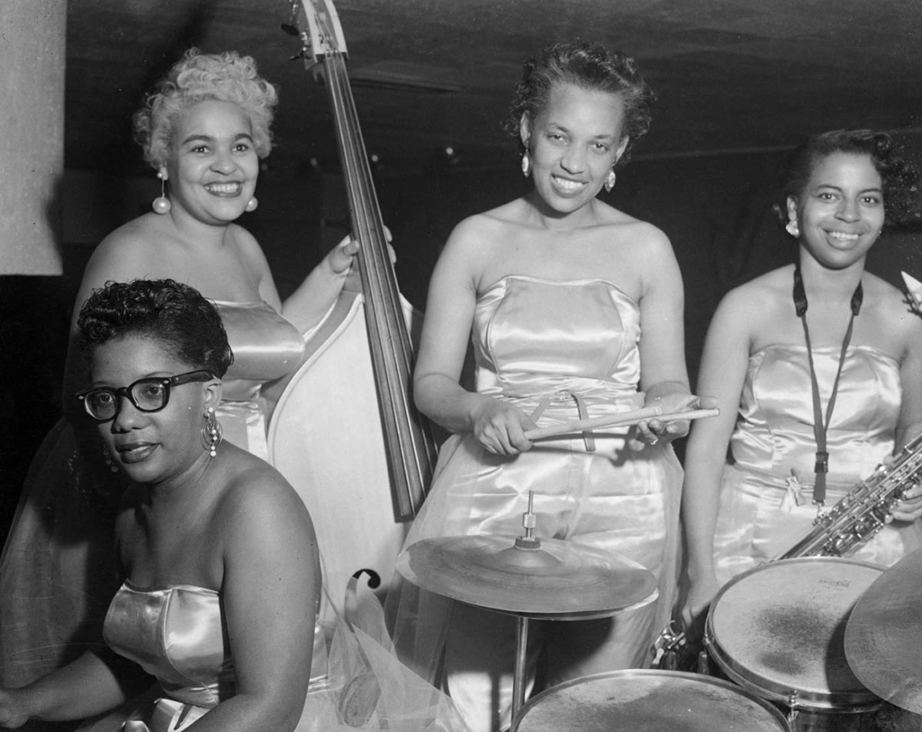 Four people in satin strapless dresses smile at the camera. One person sits at a piano, another is holding a large bass, another is holding drumsticks, and the last person is holding a saxophone