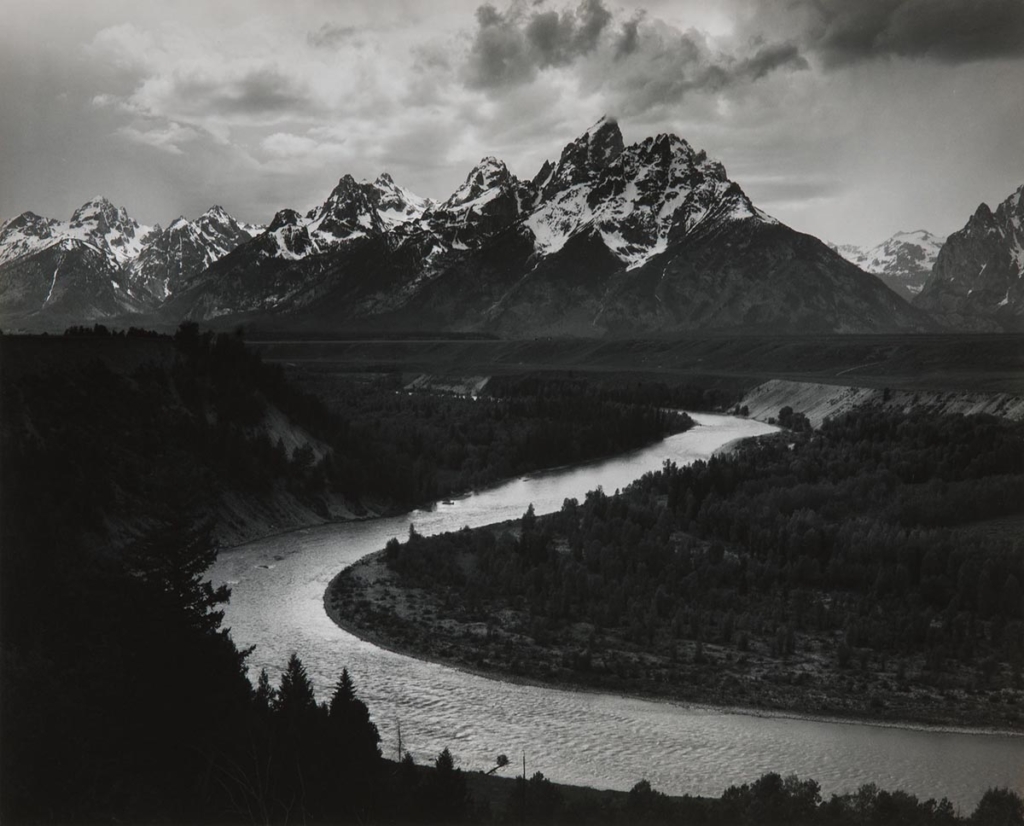 A black and white photo featuring large, snow-capped mountains with a river curving out from the mountains into the foreground