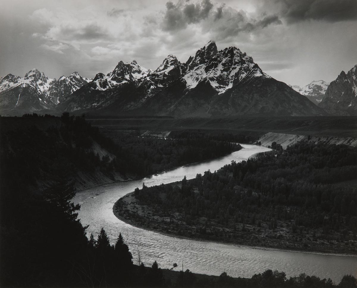 A black and white photo featuring large, snow-capped mountains with a river curving out from the mountains into the foreground