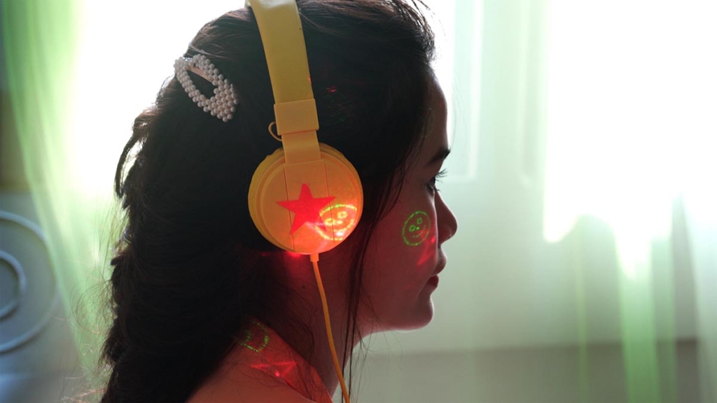 A profile portrait of a person wearing large, bright headphones and standing in front of gauzy curtains with light seeping through