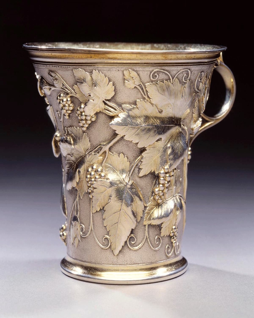 A silver and silver gilt cup covered in intricate ornamentation shaped like grape vines with a slender handle.