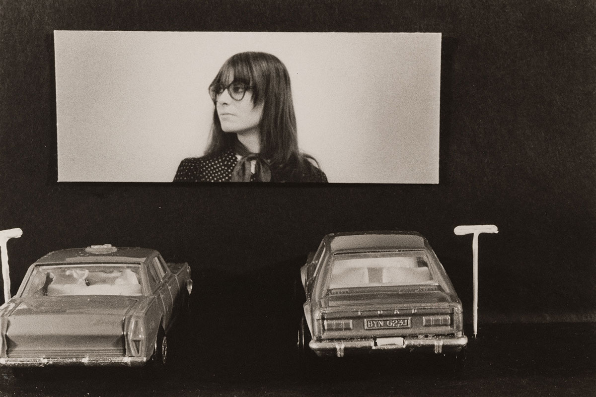 A black and white photo of two old-fashioned cars parked at a drive-in movie theater; an image of a young woman with long, dark hair and glasses is on the movie screen