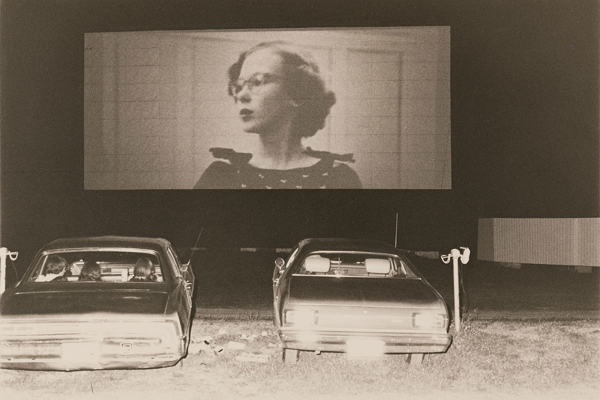 A black and white photo of two old-fashioned cars parked at a drive-in movie theater; an image of a young woman with short, curled hair and glasses is on the movie screen.