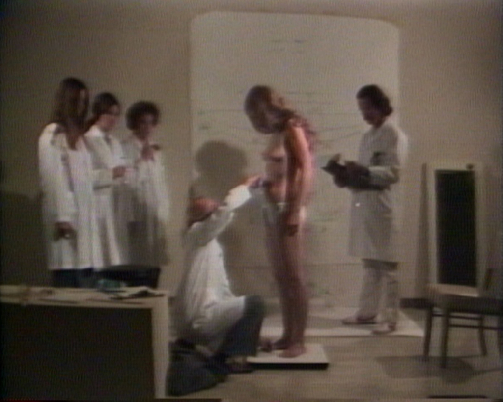 A naked woman stands on a scale surrounded by people in lab coats.