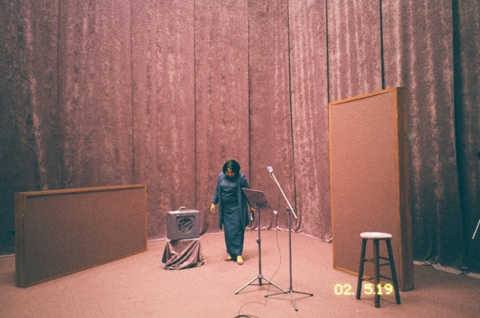 A woman stands on a stage, surrounded by recording equipment, velvet curtains hang in the background