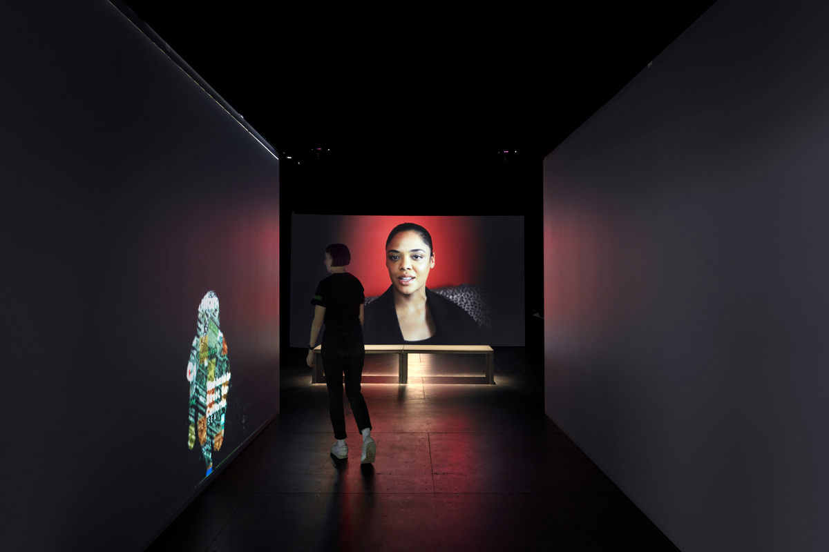 An art gallery with a video of a person playing in the background and a projected artwork displayed on the walls.