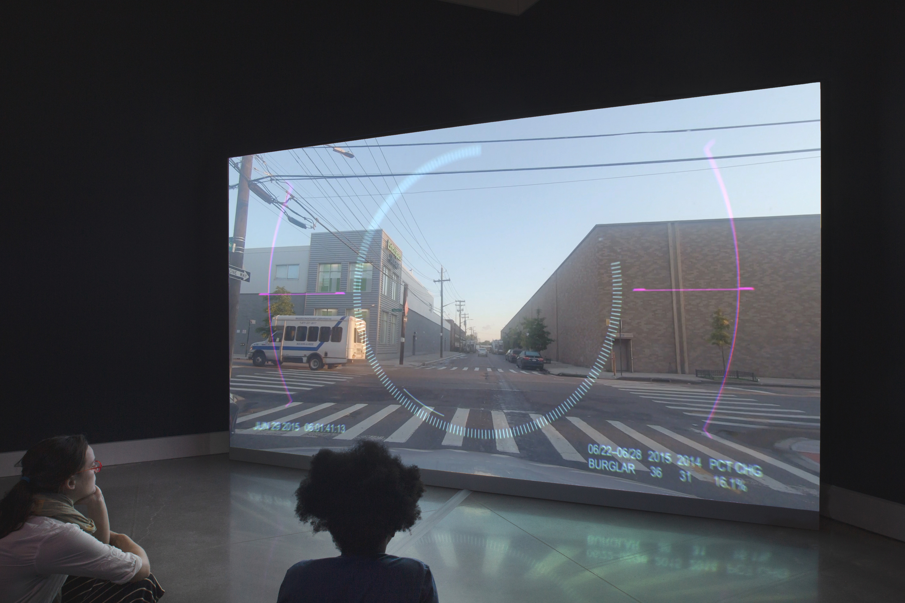 A person in a gallery looks at a video that depicts an empty street in an industrial neighborhood. Graphics that appear out a combat video game overlay the scree.