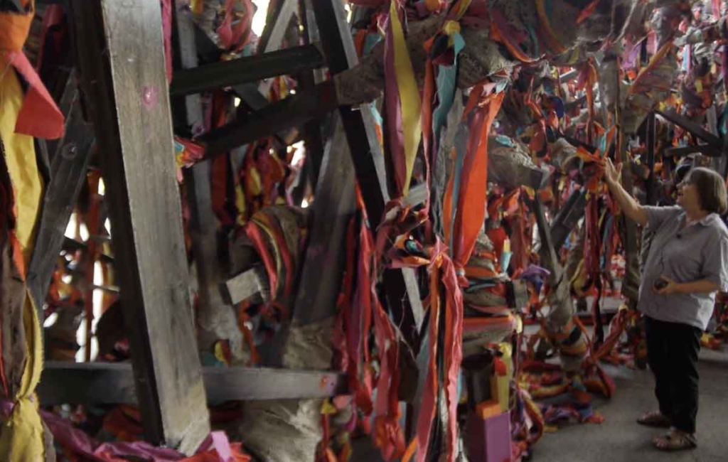 A person stands inside a large installation sculpture made from wood and large ribbons, they are touching one of the ribbons
