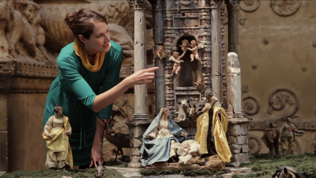 A person leans over a number of miniature figures and points at an angel suspended in the air.