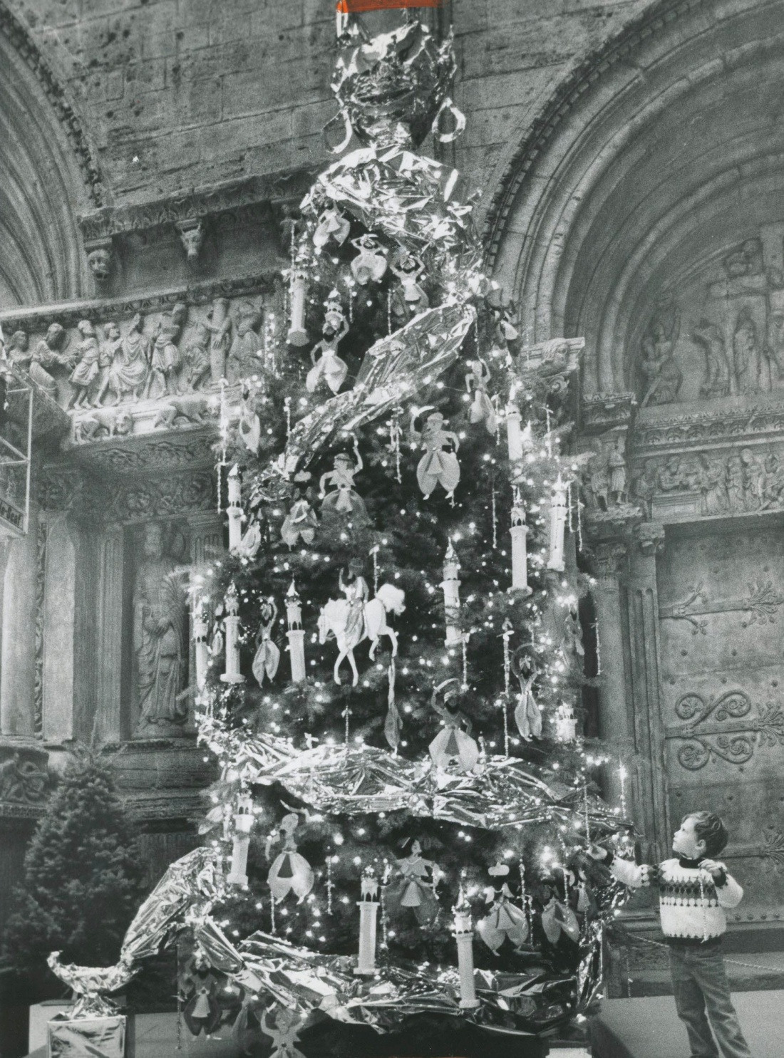 A black and white photo of a small child standing by alarge decorated Christmas tree.