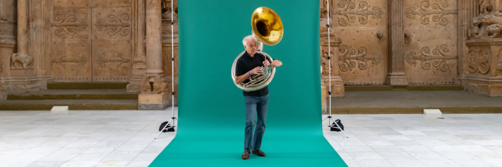 A man and a brass instrument stand on a green screen.