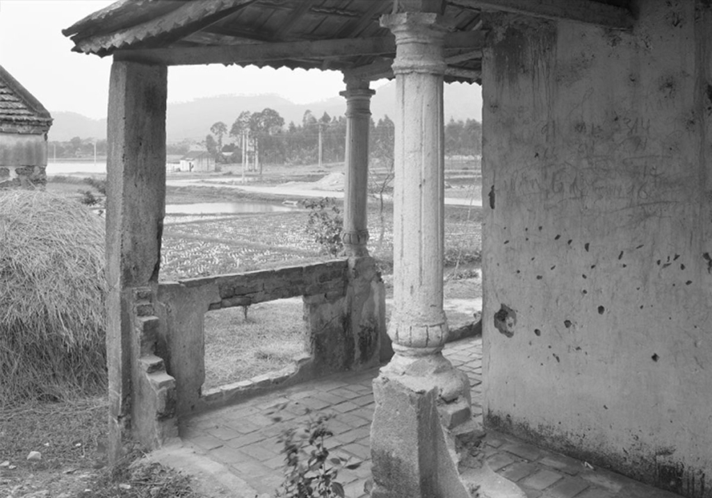 A black and white photo of the corner of a building with a roof held up by columns; a wide landscape with trees in the distance is beyond the open porch