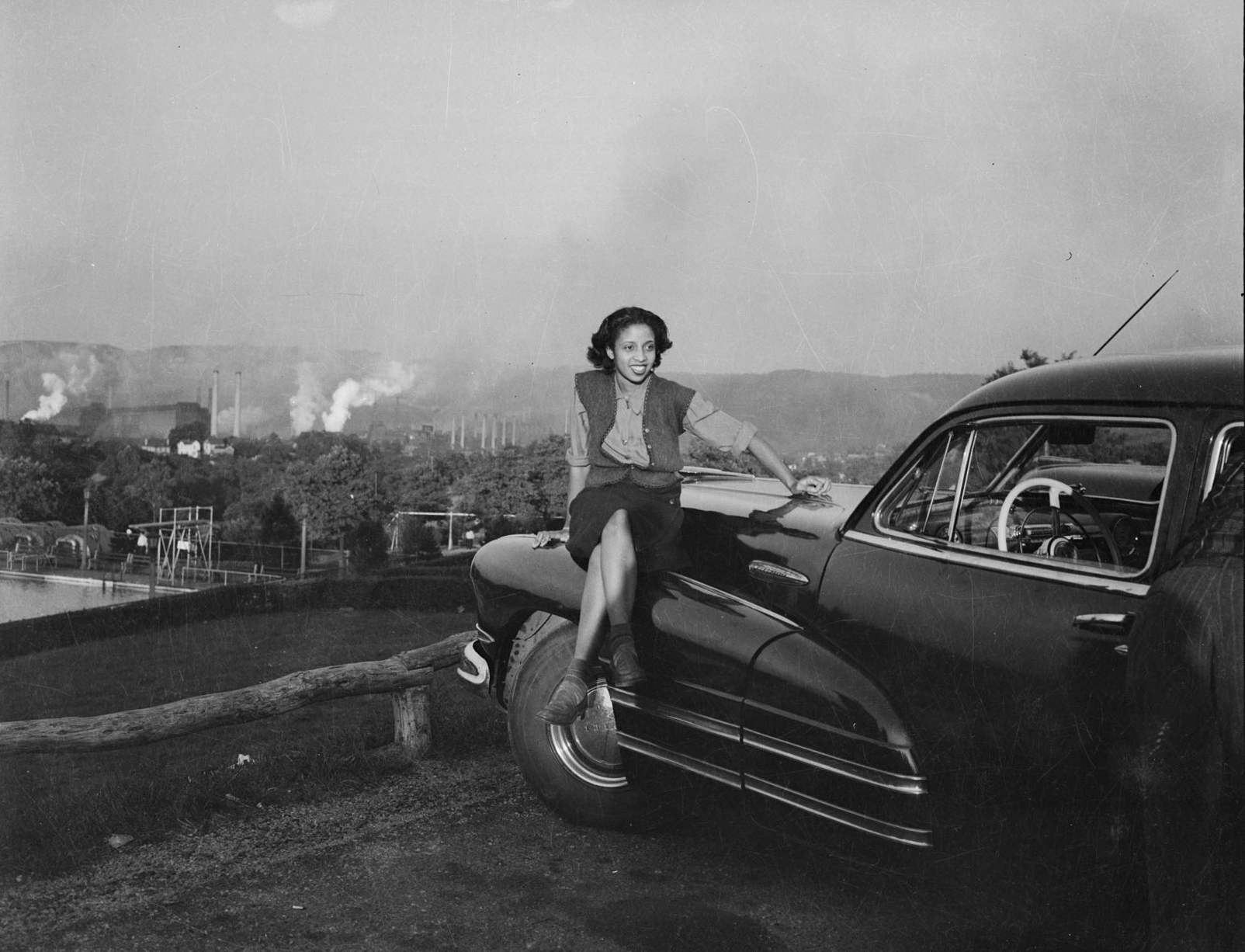 Black and white photograph of woman seated on hood of a Buick with steel mill in background.