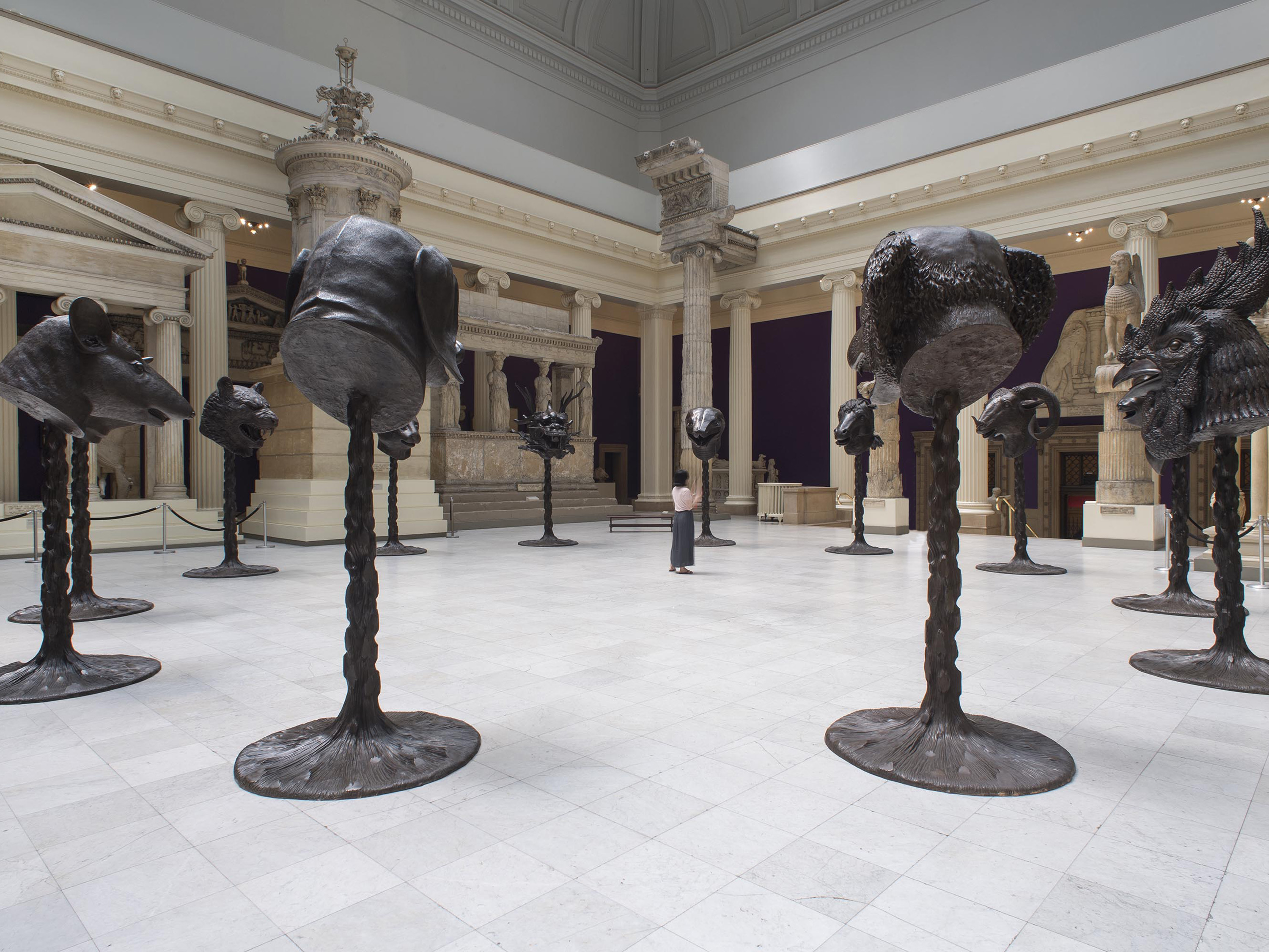 A large gallery room with five bronze animal head sculptures presented on tall, narrow bronze posts in front of an architectural cast of an ornate doorway with columns