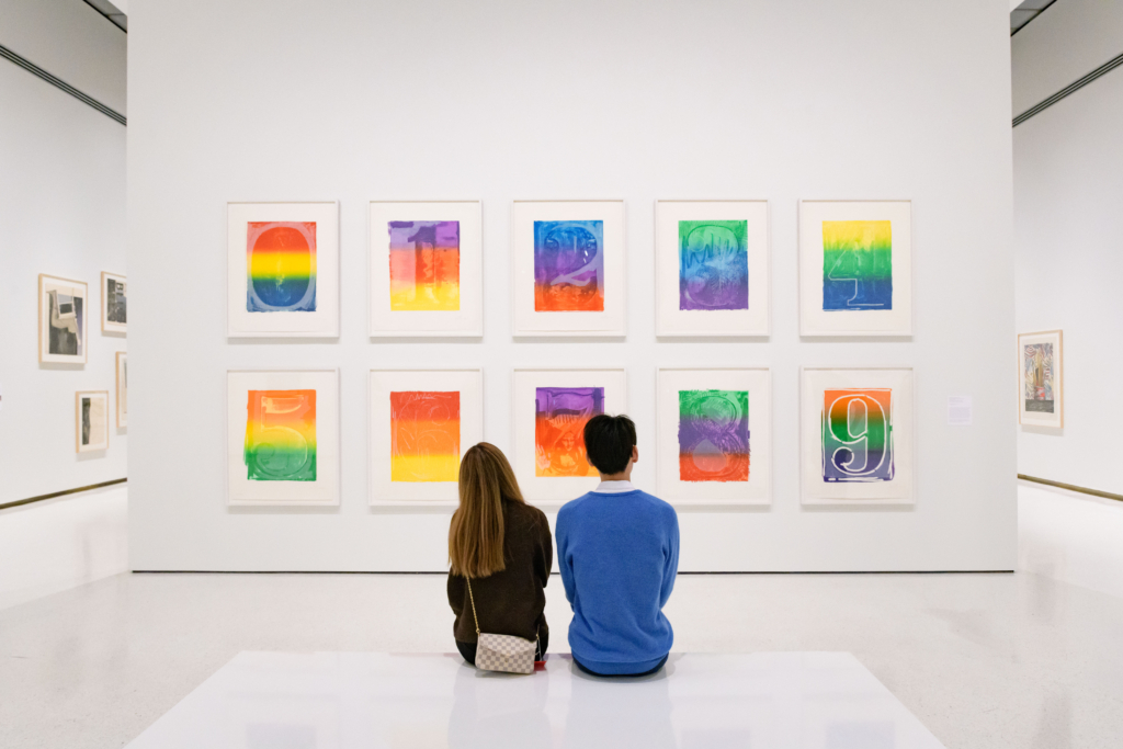 Two people sit in a gallery, looking at a wall of ten colorful prints showing each numeral up to 10.