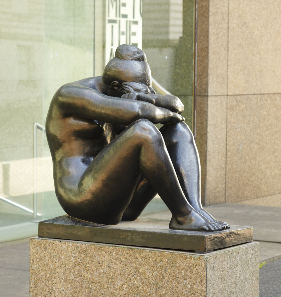 A sculpture depicts a woman seated with her knees drawn up. She is resting her elbows on her knees and her head on her forearms and her hair is in a bun.