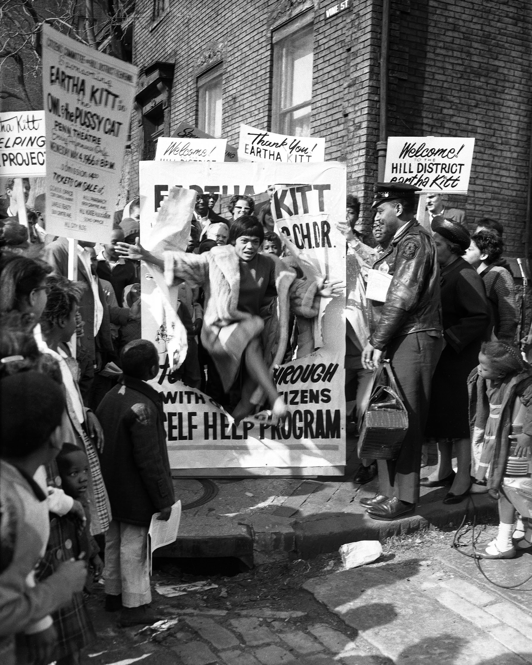 Charles Teenie Harris photo of Eartha Kitt leaping though poster to launch self help drive by Citizens Committee on Hill District Renewal, surrounded by crowd, including police officer Harvey Adams, Vine and Colwell Streets Hill District