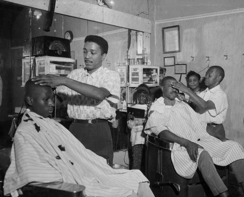 Charles Teenie Harris Barbers Pete Boyd and Johnny Gator cutting man’s and boy’s hair with woman and girl looking on, in Johnny Gator’s barbershop