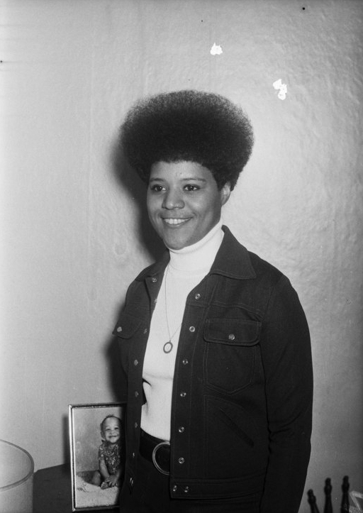 Charles Teenie Harris Woman wearing afro, round pendant, and jacket with snap closures, standing in interior with portrait of baby in background