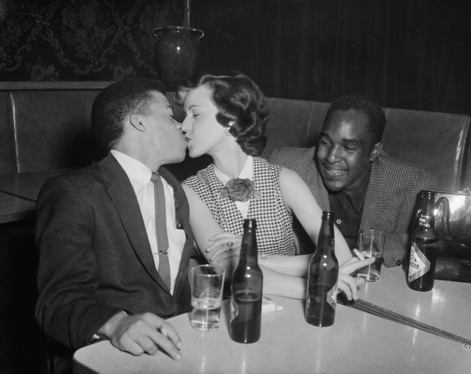 Man and woman kissing, with smiling man looking at them, behind table with bottles of Duquesne Beer, in bar or restaurant, ca. 1950–1965